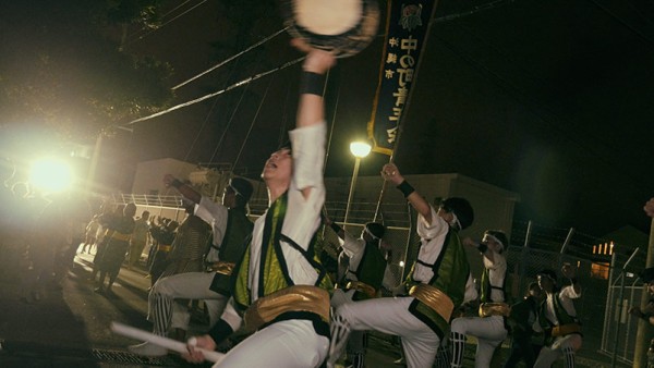 a drummer raises the small drum high and male dancers on the back
